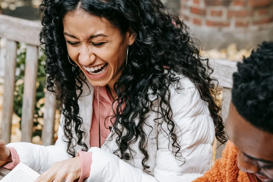 Two students laughing over an open book on a bench outside